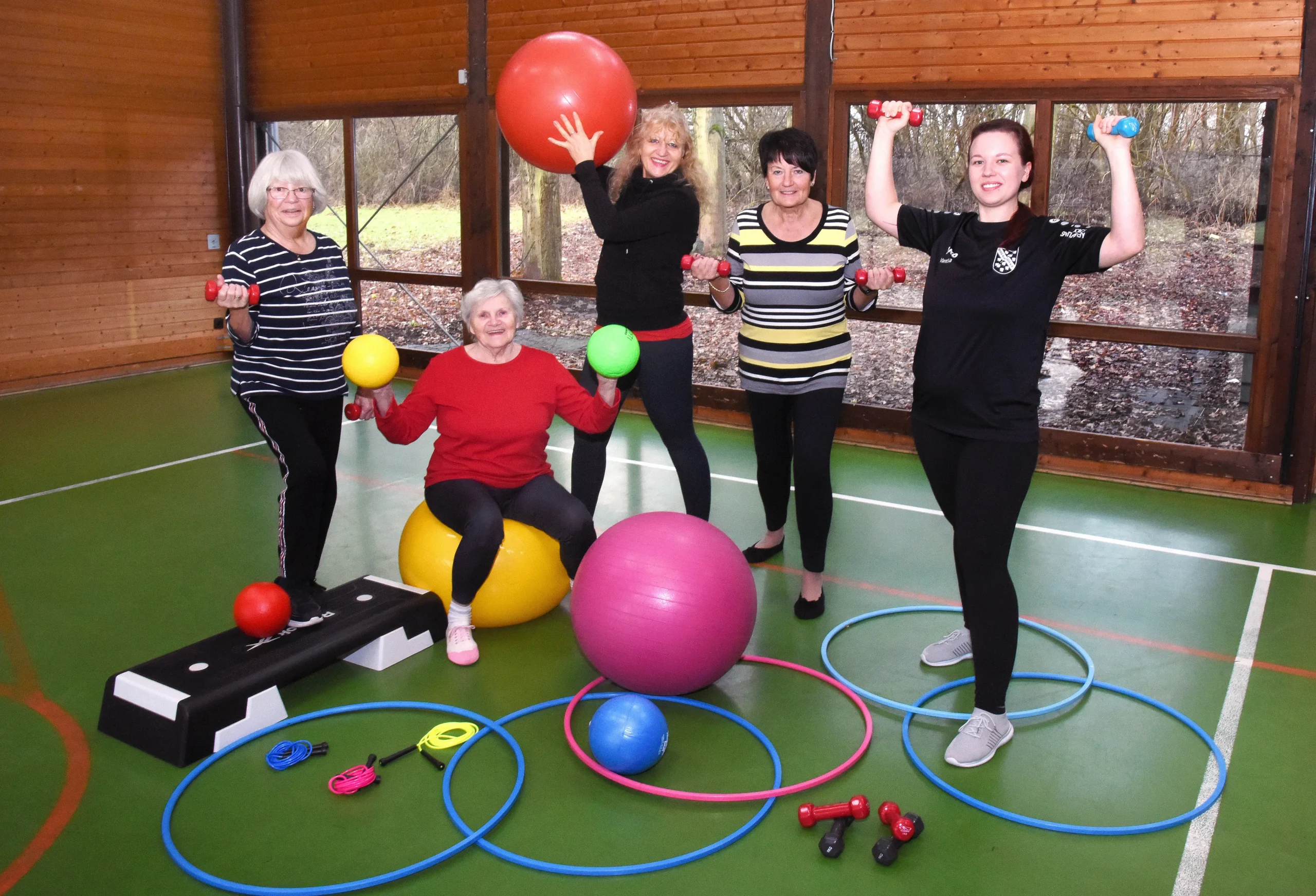 Fünf Frauen unterschiedlichen Alters posieren fröhlich in einer Sporthalle mit Fitnessbällen, Hanteln, Reifen und Springseilen. Sie strahlen Energie und Freude an Bewegung aus.
