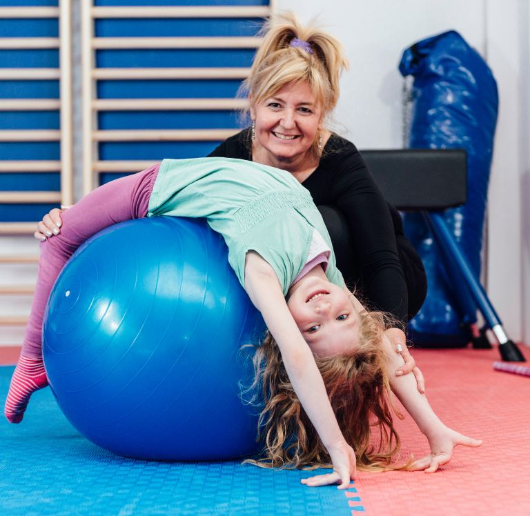 Eine Frau hilft einem Kind, das auf einem blauen Gymnastikball liegt, bei einer Dehnübung. Beide lächeln in die Kamera.