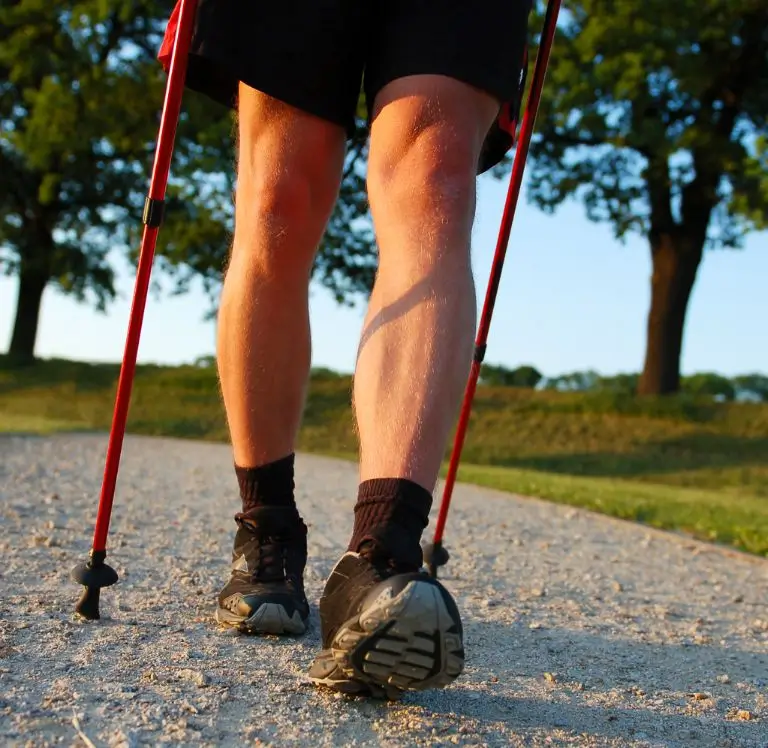 Nahaufnahme von Beinen einer Person, die mit Wanderstöcken auf einem Schotterweg spaziert. Bäume und Natur im Hintergrund.