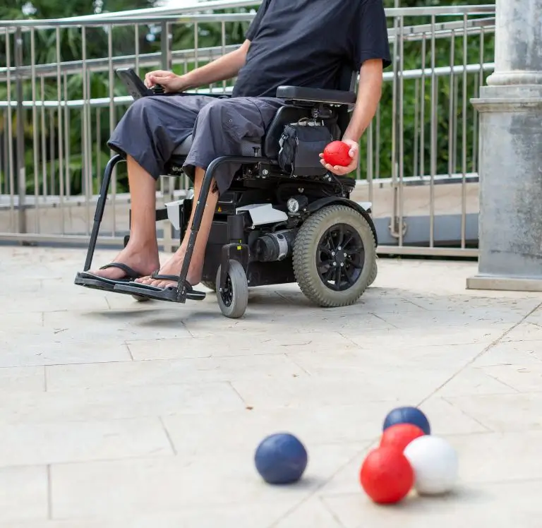 Person im Elektrorollstuhl hält eine rote Boccia-Kugel, während mehrere Boccia-Kugeln auf dem Boden verteilt liegen.