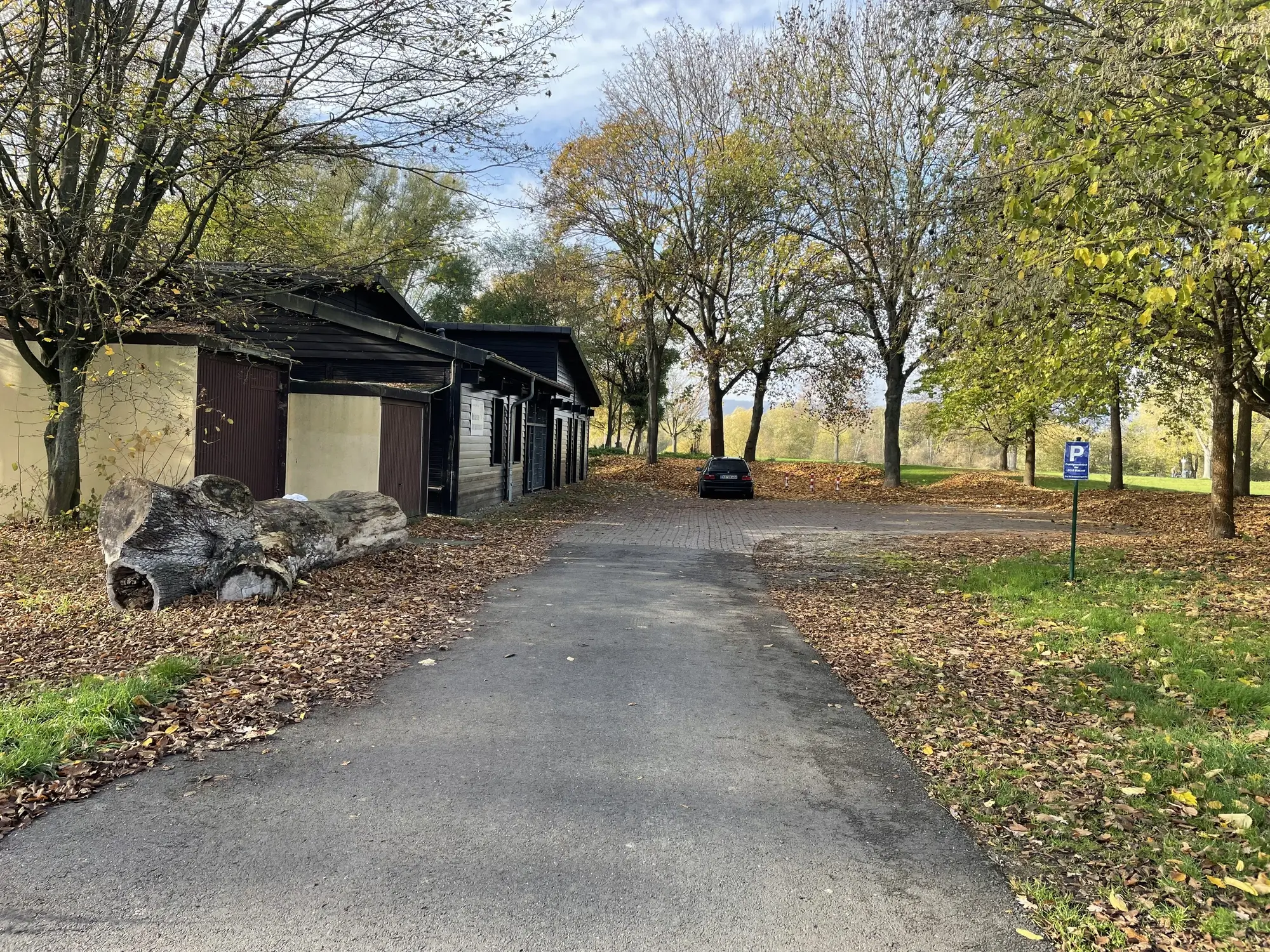 Vereinsheim in herbstlicher Umgebung, umgeben von Bäumen mit fallendem Laub. Ein Parkplatz mit einem Behindertenparkplatzschild ist rechts zu sehen